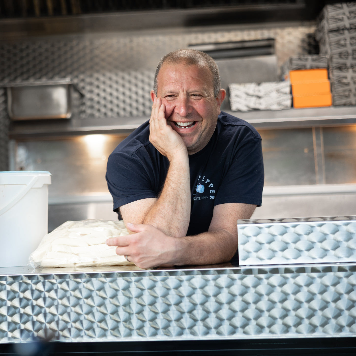 Two men in a kitchen setting, one leaning on a counter with a pizza, smiling.