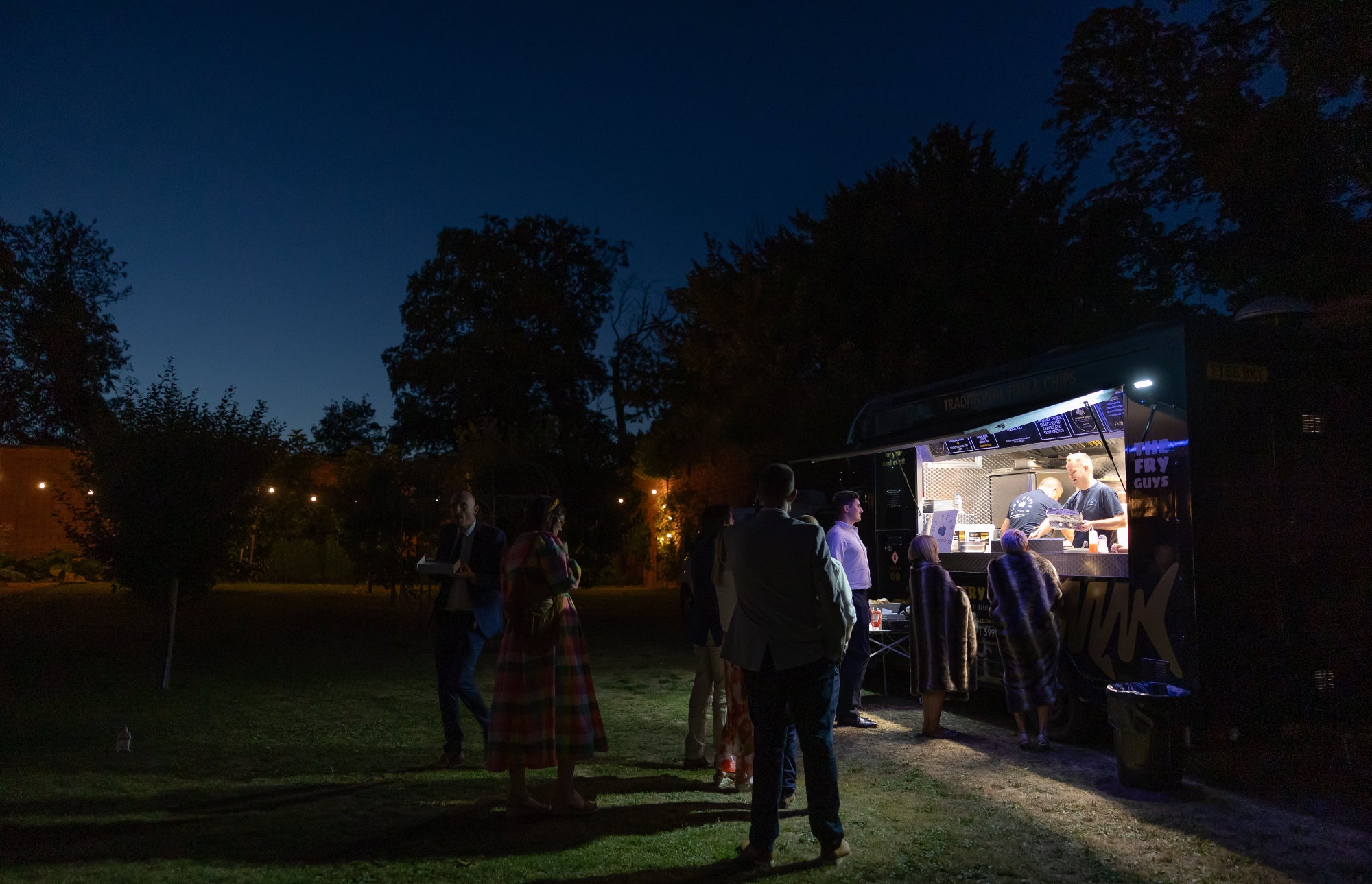 People gathered around a food truck at night with trees in the background